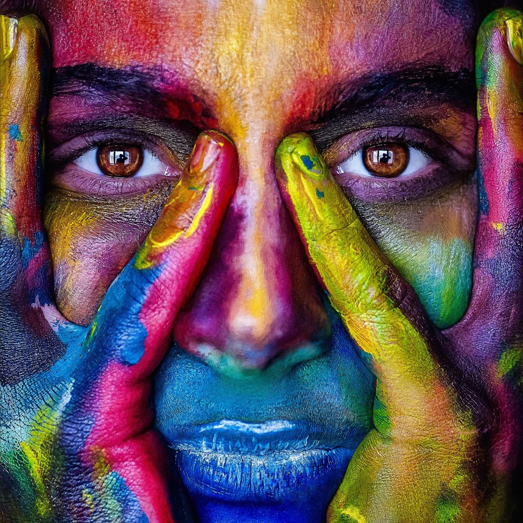 Woman covered in multi-coloured paint staring at the camera, hands against her cheeks.